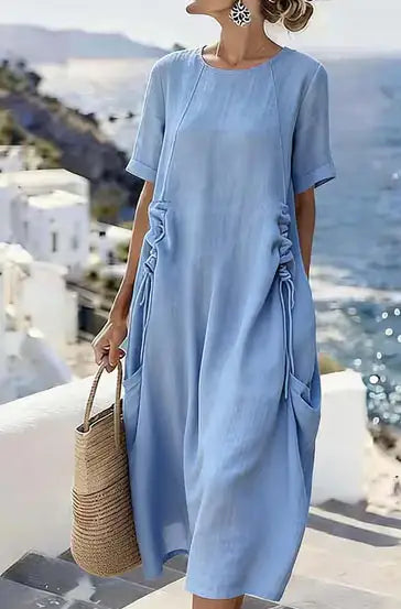 Woman wearing a light blue dress with a straw bag, standing by a scenic coastal view.